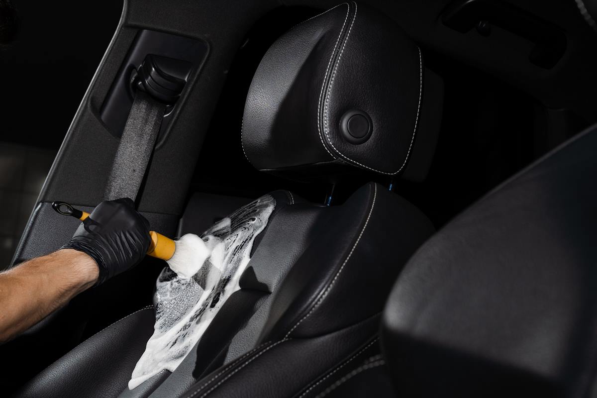 Behandschuhte Hand tr&auml;gt mit der B&uuml;rste Schaum auf einen schwarzen Ledersitz auf